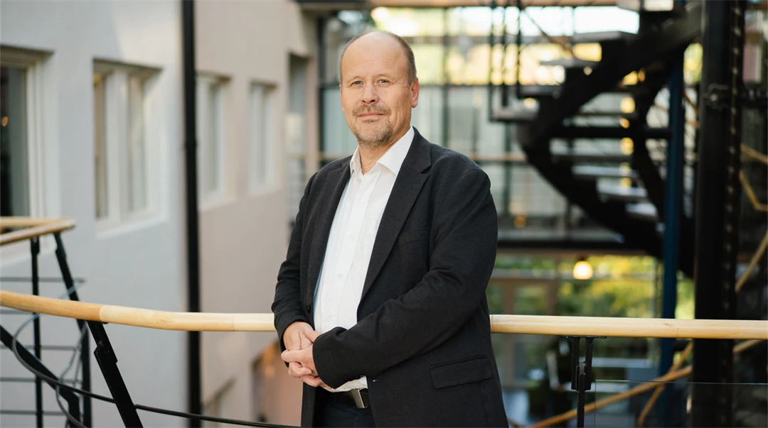 Man i vit skjorta och svart kavaj med knäppta händer tittar in i kameran. 