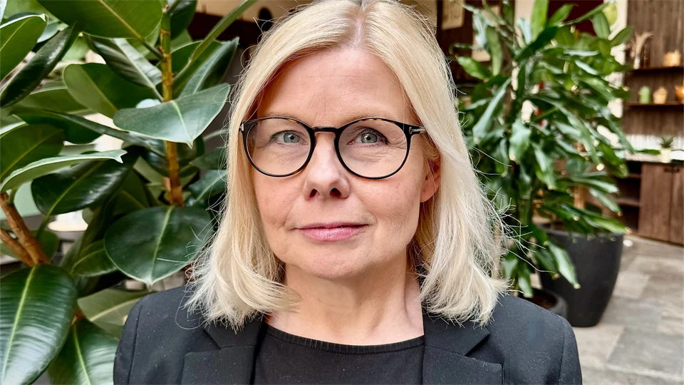 En kvinna med axellångt blont hår och glasögon, klädd i svart kavaj står framför gröna, höga växter i inomhusmiljö. 