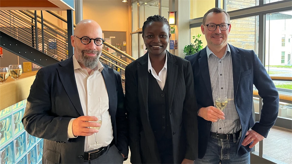 Tre personer, två män och en kvinna i kostymer tittar in i kameran och ler. Männen håller i varsitt glas med ciderdryck. 