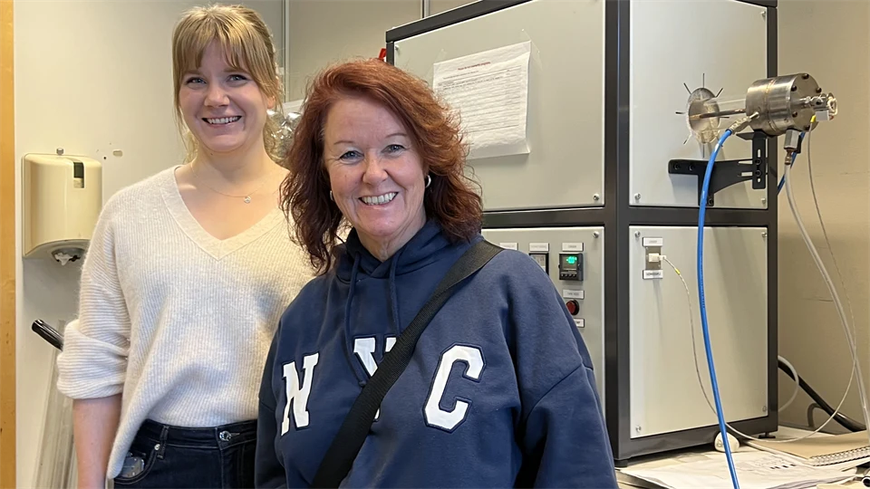 Två kvinnor står bredvid varandra i ett rum framför en ljusgrå låda med knappar på och en blå slang.