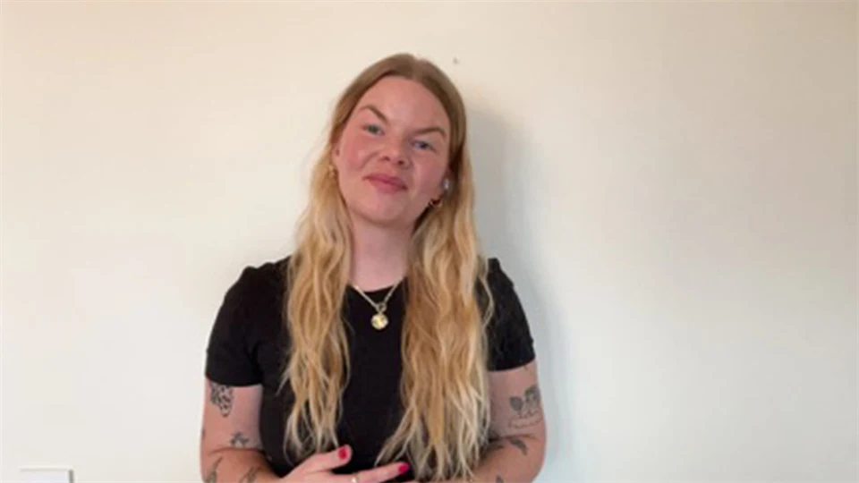 Kvinna i svart kortärmad tröja och långt blont hår står framför en beige vägg. 