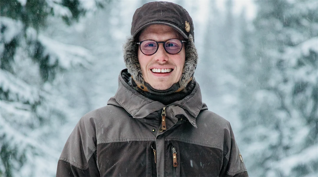 Glad ung man i tjocka vinterkläder står utomhus i vinterlandskap med snöklädda granar i bakgrunden