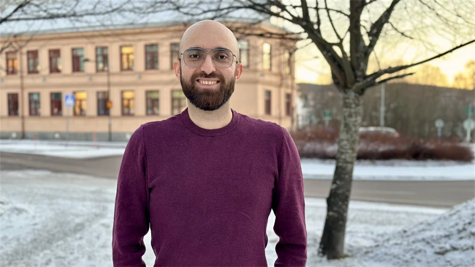 Smiling man with a shaved head, brown beard, and glasses. He is wearing a burgundy sweater and standing outdoors. The season is winter.