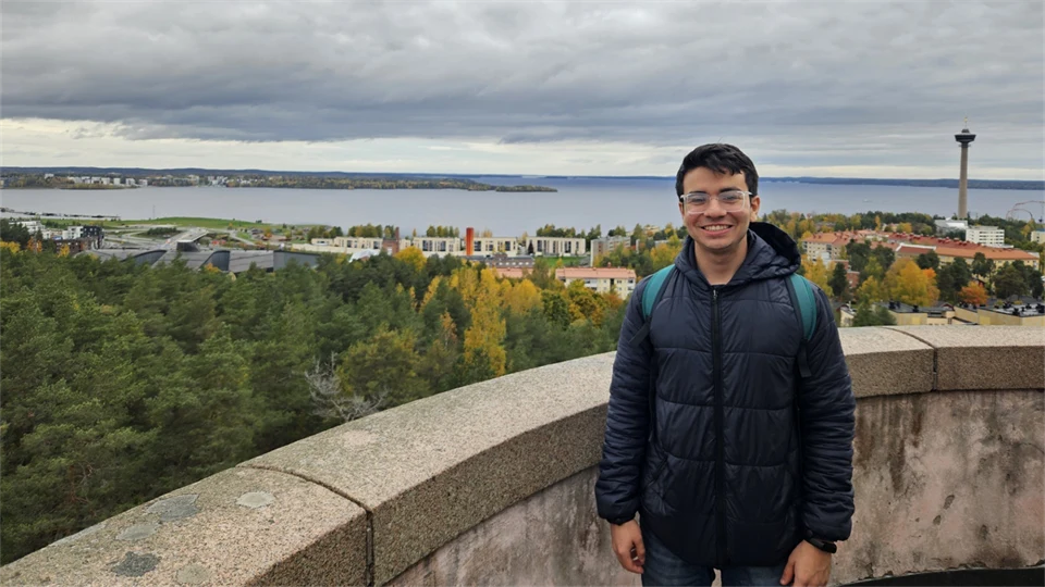A young man wearing a dark blue jacket and carrying a rucksack is standing by a stone wall high up, looking out over trees, buildings and water. 