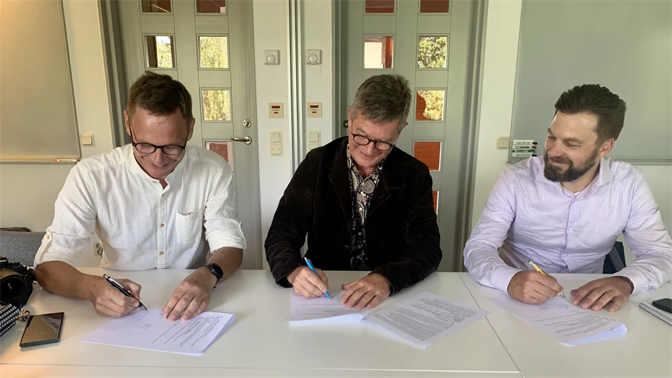 Tre män sitter vid ett bord och signerar ett avtalsdokument