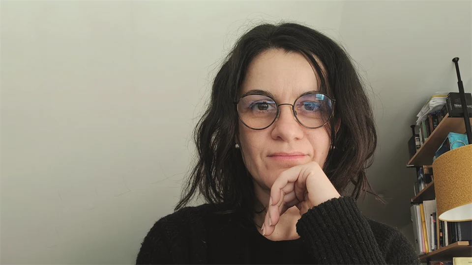 En mörkhårig kvinna med glasögon vilar hakan mot sin hand och tittar in i kameran.