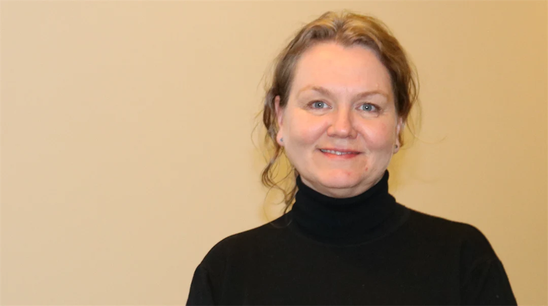 A woman with brown hair tied up and wearing a black polo shirt in a corridor with beige walls.