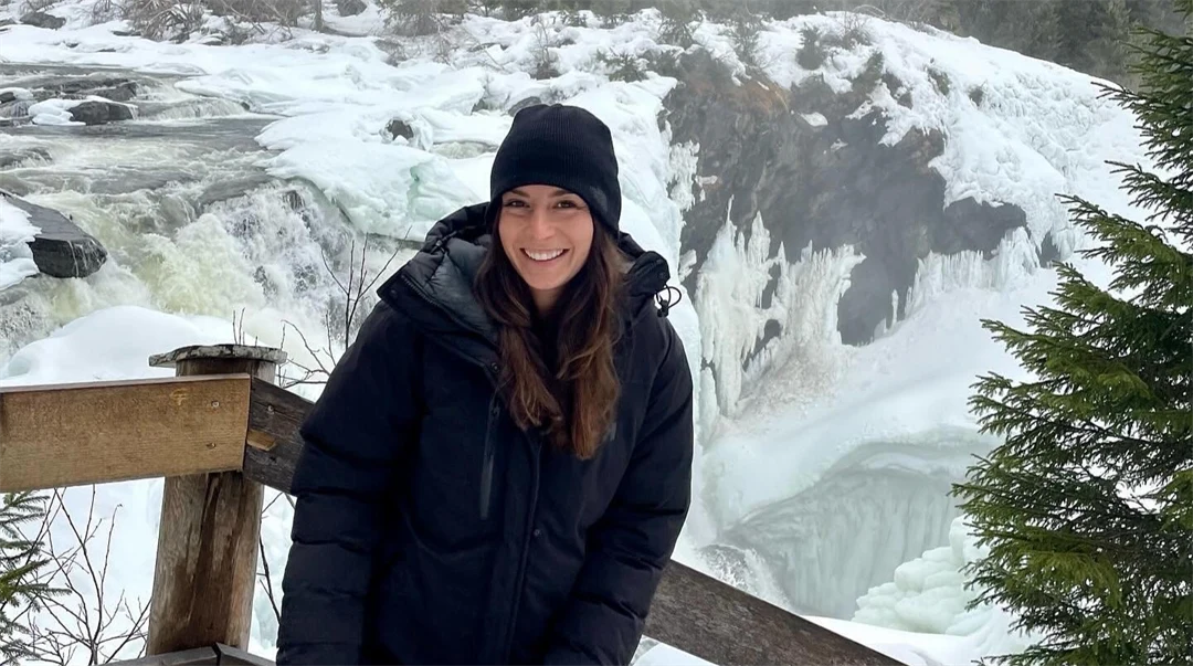 Young woman in black cap and black jacket standing in front of a waterfall