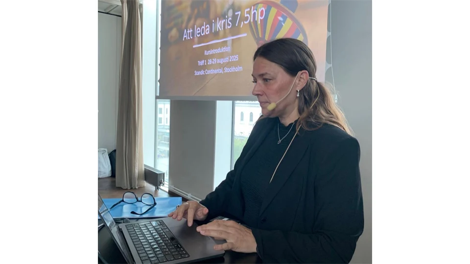 En kvinna med mikrofon står vid en bärbar dator med en skärm snett bakom sig. 