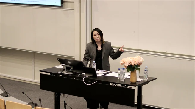 Person står vid ett bord, pekar mot en skärm och presenterar material samtidigt som personen talar till en publik.