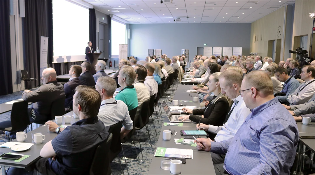 A large group of people are gathered at a conference and are listening to a speaker showing images on a large screen.