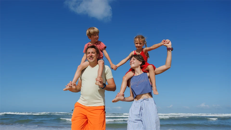 Mamma och pappa bärandes varsitt barn på axlarna. De befinner sig på en soldränkt strand. 