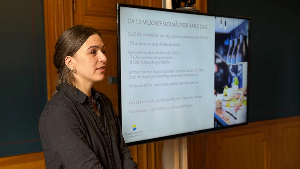 En kvinna i brunt hår och mörkgrå skjorta står vid en skärm med en presentation på. 