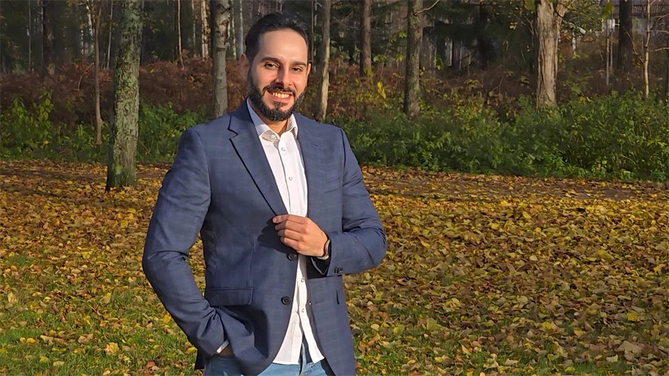 A man looks into the camera and smiles, in the background there are trees and yellow leaves on the ground.