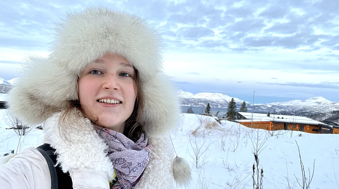 A young woman wearing a white fur hat and white winter jacket with a fur collar stands in a winter landscape with mountains, sea and a building in the background.