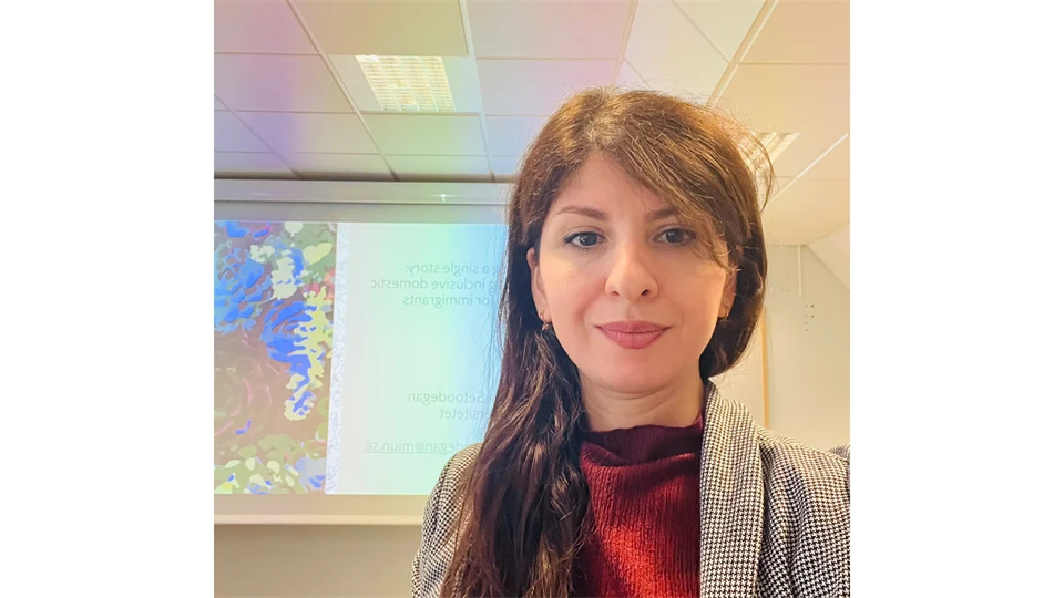 Kvinna i mörkt långt hår, grå kofta och röd blus står framför en skärm med en presentation på.