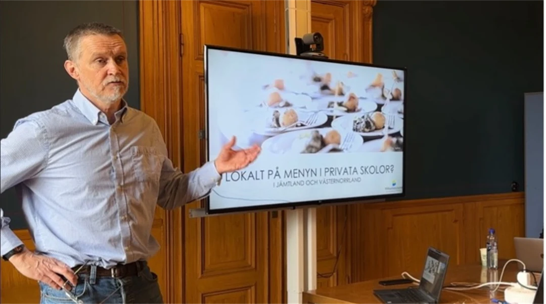 En man i ljusblå skjorta står framför en skärm med en presentation på och gestikulerar med sin vänstra hand.