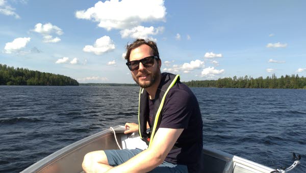 En man med solglasögon och marinblå t-shirt sitter i en båt, med sjön och skog i bakgrunden. 