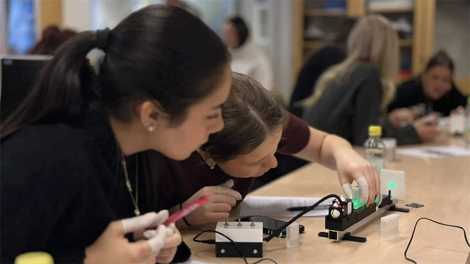 Två kvinnor sitter vid ett bord och undersöker en utrustning. I bakgrunden syns fler personer. 