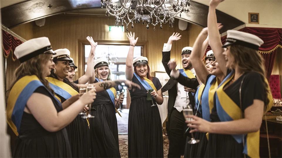 A group of happy young people wearing student hats cheer with one hand in the air.