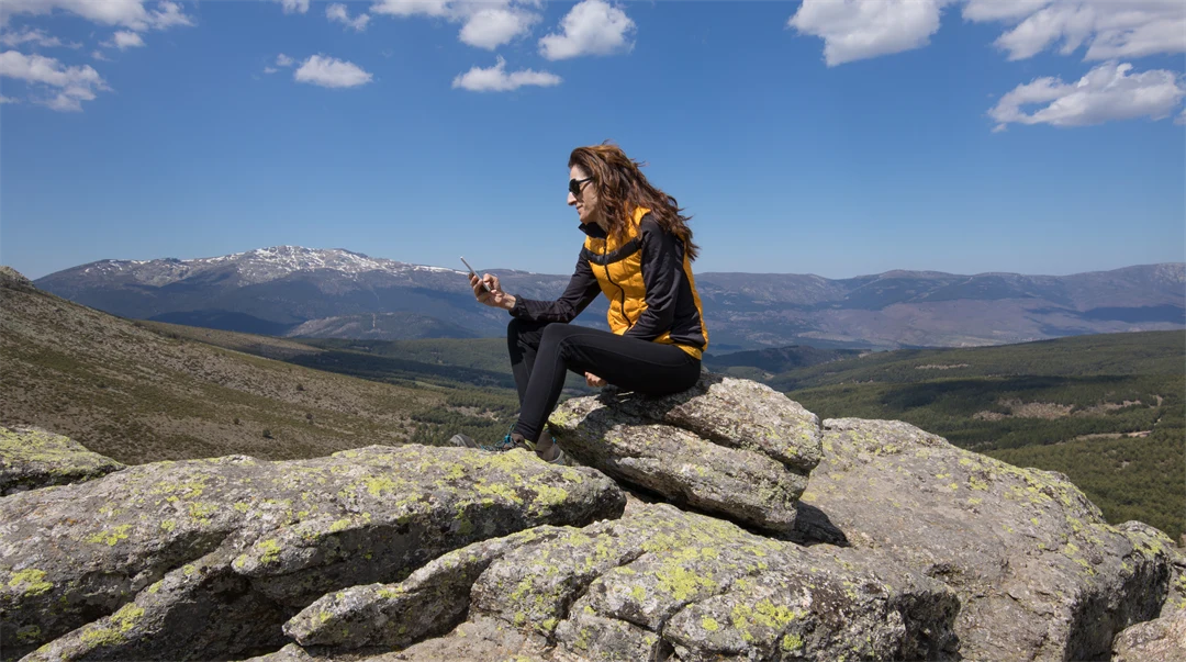 sport hiking or trekking woman with yellow and black clothes, sitting on rock peak, typing mobile smart phone, behind large mountains 