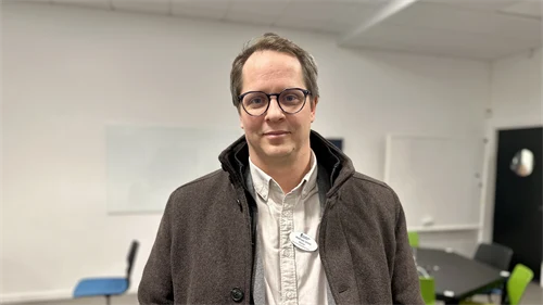 En man i brun jacka och vit skjorta står i klassrum med färgglada snurrstolar i bakgrunden.