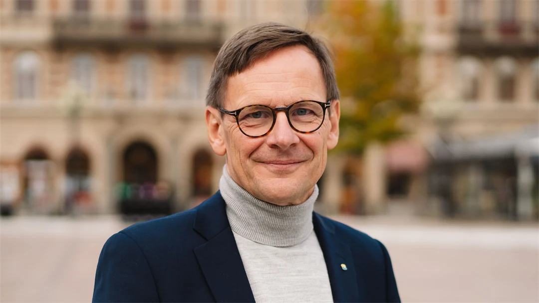 En mörkhårig man med glasögon, grå polotröja och mörk kavaj står på ett torg och tittar in i kameran. 