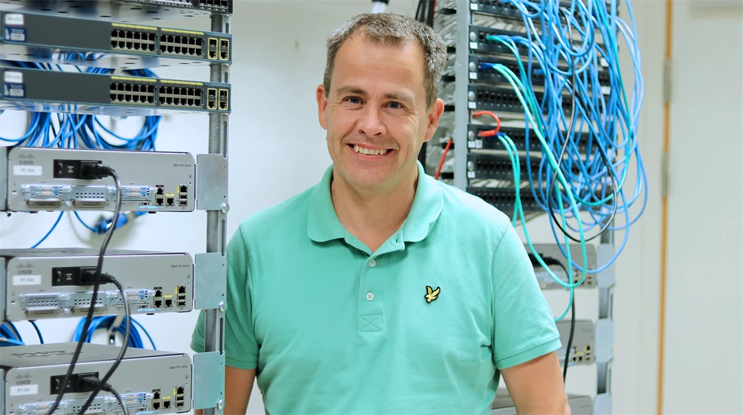 Man i turkos pikétröja framför teknikskåp med sladdar