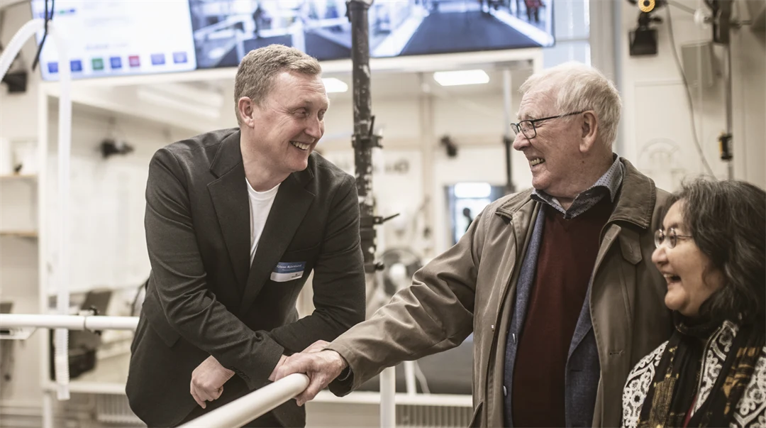 Man står lutandes mot staket och ler mot glad kvinna och glad man i ett forskningslabb. 