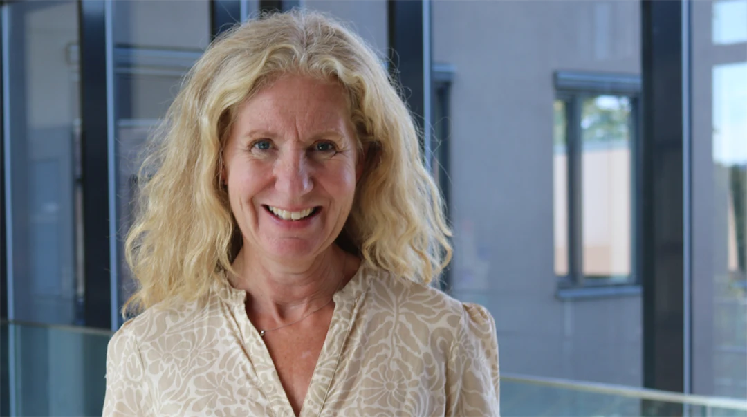 A woman with light hair and a cream-coloured blouse stands in a glass corridor with windows behind her.