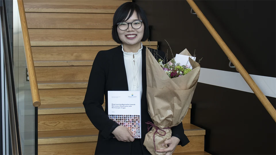 Yali Nie poses in front of the camera with her thesis and a bouquet of flowers.