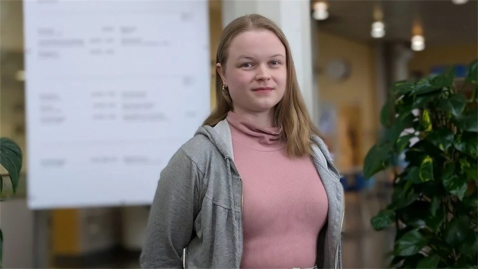 En kvinna med långt hår står framför en ljustavla. Bredvid finns en grön växt.