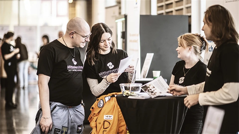 Two male and two female students in black Mid Sweden University t-shirts stand around a table reading on a postcard.