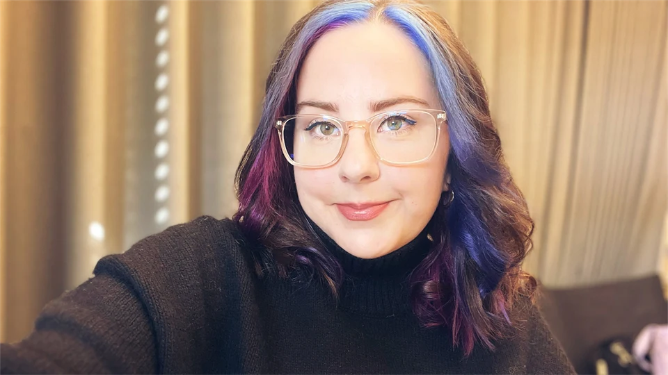 En kvinna med glasögon och mörkt hår med lila slingor sitter framför ett draperi