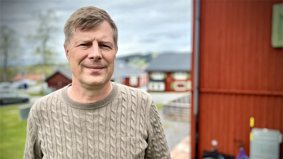 Man i beige tröja står utomhus med en röd husvägg i bakgrunden. 