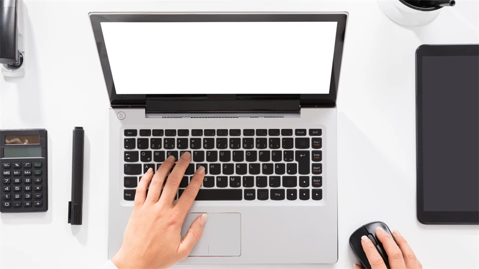 Händer knappar på en laptop, miniräknare, tuschpenna och häftmaskin ligger in till