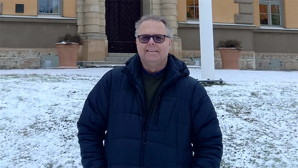 En man i mörk jacka och glasögon står utomhus.