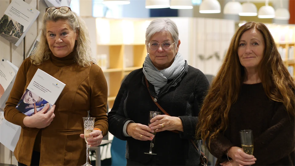 Tre kvinnor står bredvid varandra med champagneglas i händerna. Kvinnan längst till vänster håller upp en bok.