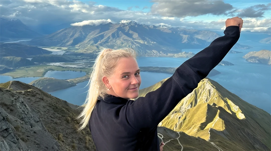 Ung kvinna står på ett berg med utsikt över havet och ett vulkanlandskap. Hon är glad och sträcker upp sin arm. 