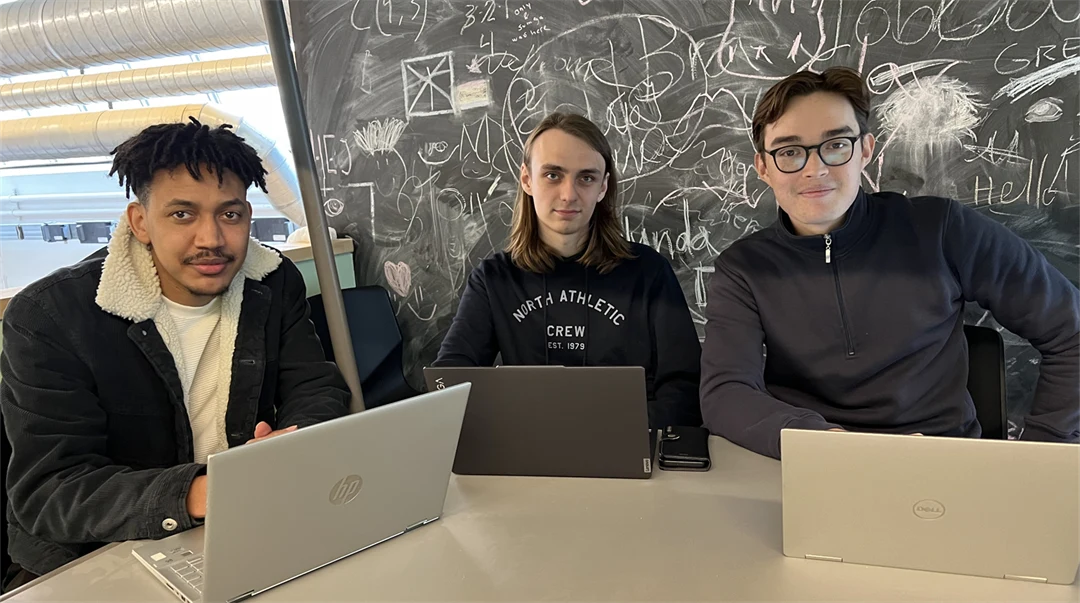 Three young men sit together at a table with their laptops in front of them.