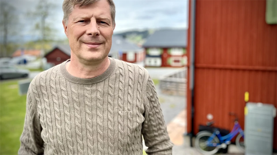 Man i beige tröja står utomhus med en röd husvägg i bakgrunden. 