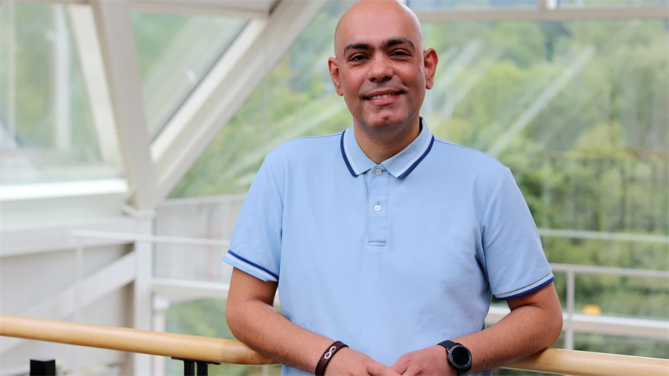 Man in blue short-sleeved shirt stands by a railing and smiles at the camera.