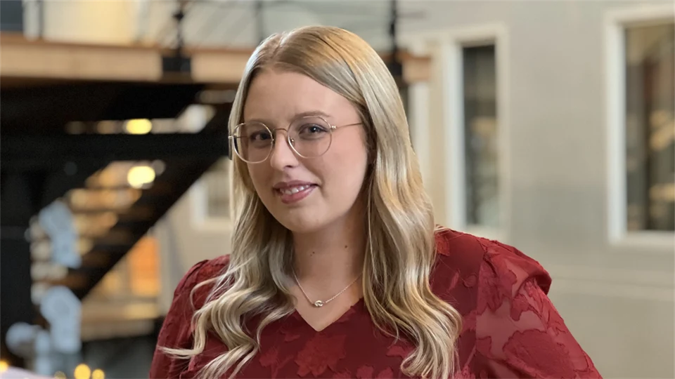 A young woman with long hair and glasses looks into the camera.