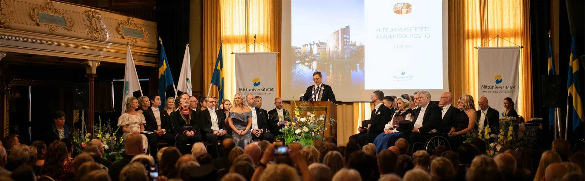 En finklädd man står i en talarstol och talar framför en publik.