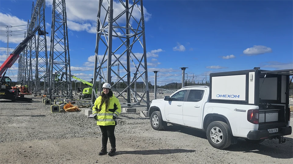 En kvinna i gul jacka och vit hjälm står på en grusplan, bredvid en vit bil. I bakgrunden syns stålkonstruktioner och delar av en kranbil.