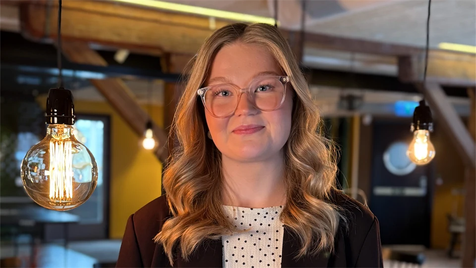 En kvinna ser in i kameran och ler. Lampor hänger i bakgrunden.