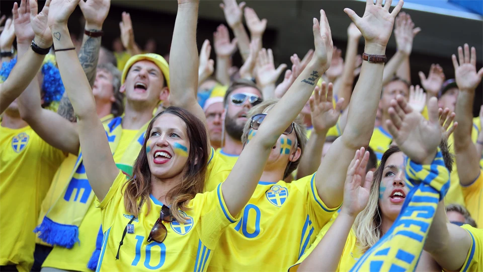 Glada människor klädda i fotbollströjor och halsdukar hejandes på Sverige på en arena. Vissa har händerna uppsträckta och vissa klappar händerna. 