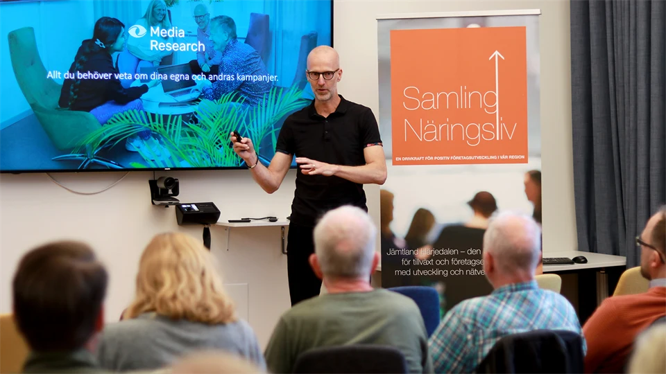 En man som står bredvid en stor skärm och talar för en sittande publik