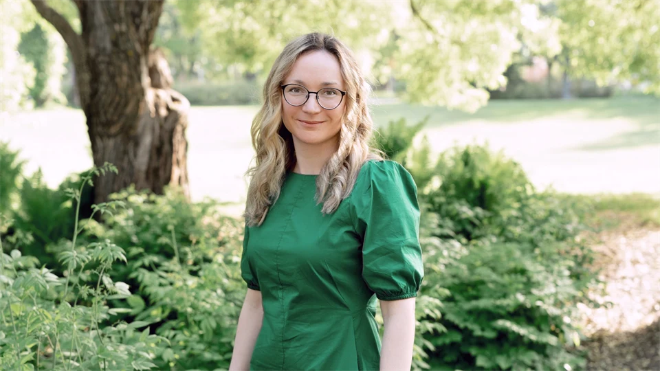 Ung kvinna med mellanlångt ljust hår och glasögon. Hon bär en grön klänning och står i en grönskande trädgård.