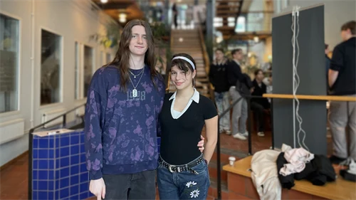 Två personer står tätt tillsammans och tittar in i kameran, människor rör sig i bakgrunden.